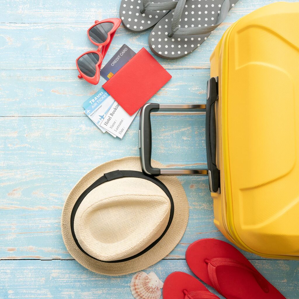 Yellow suitcase on the ground next to two pairs of flip flops, plane tickets, sunglasses and a hat.